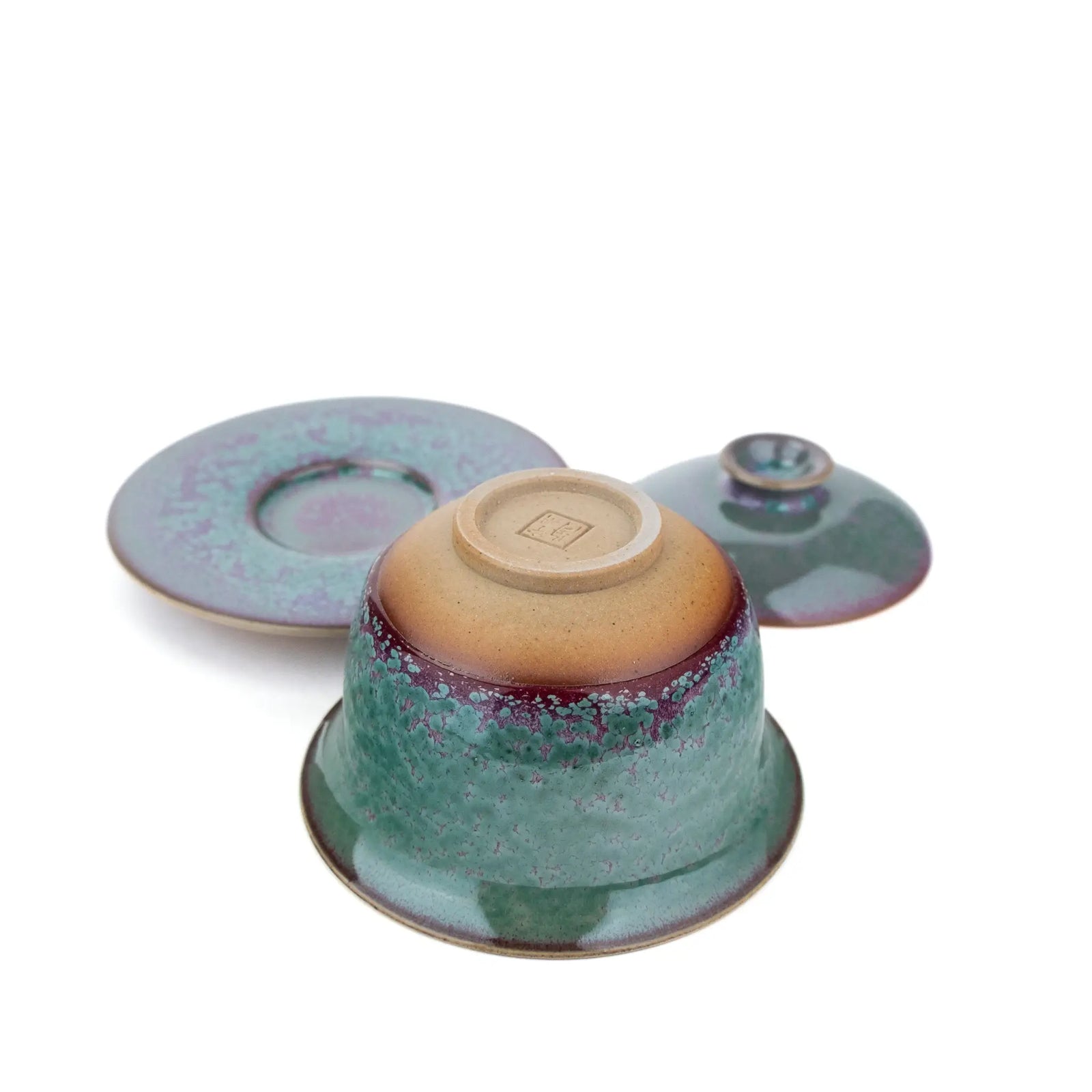 A handmade gaiwan with a matching saucer, featuring a speckled teal and purple glaze against a plain white background - underside view