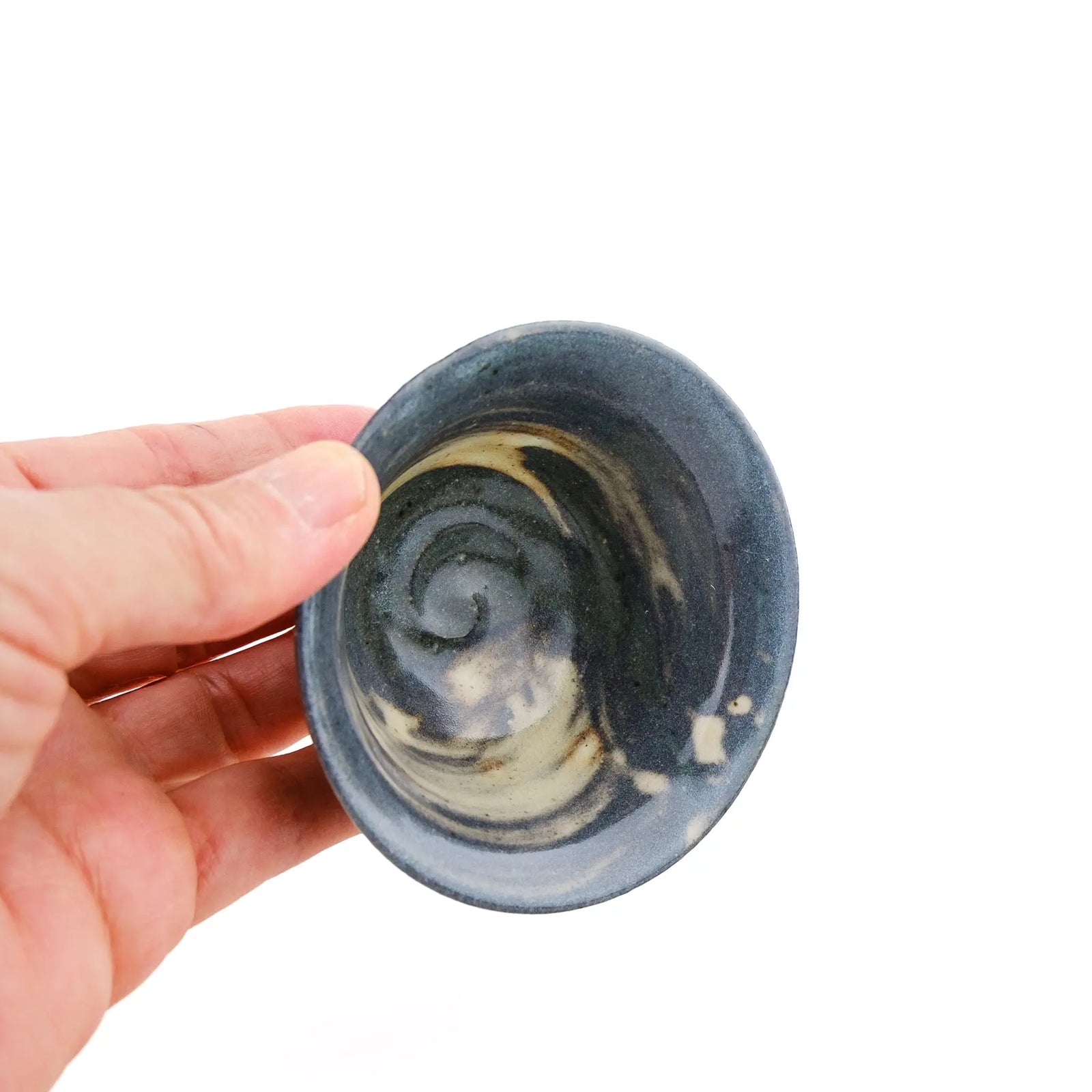 A hand holding a small handmade ceramic tea cup with a dark blue-gray exterior and a swirled cream interior glaze, shown against a plain white background.