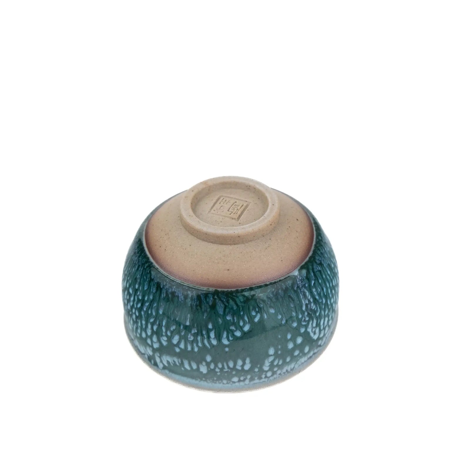 A small ceramic tea cup with a speckled dark teal-green glaze and a natural beige clay interior, photographed from above on a plain white background.