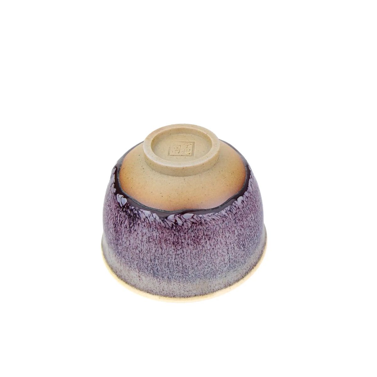 A ceramic tea cup with a purple and blue speckled glaze that fades to a beige rim and base, shown on a plain white background - Underside view
