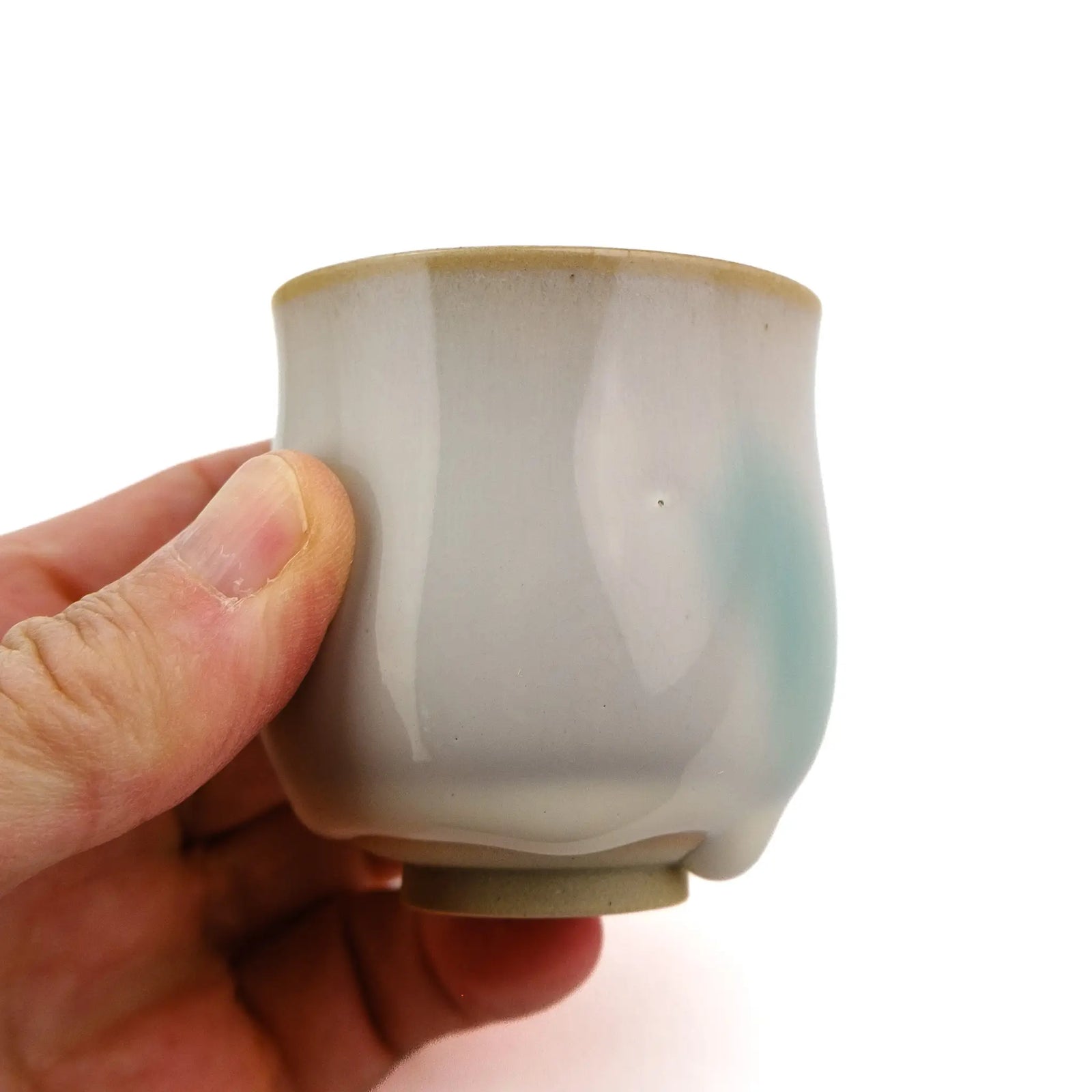 A hand holding a small ceramic tea cup with a glossy off-white glaze, featuring a light blue wash on the side and a warm beige rim.