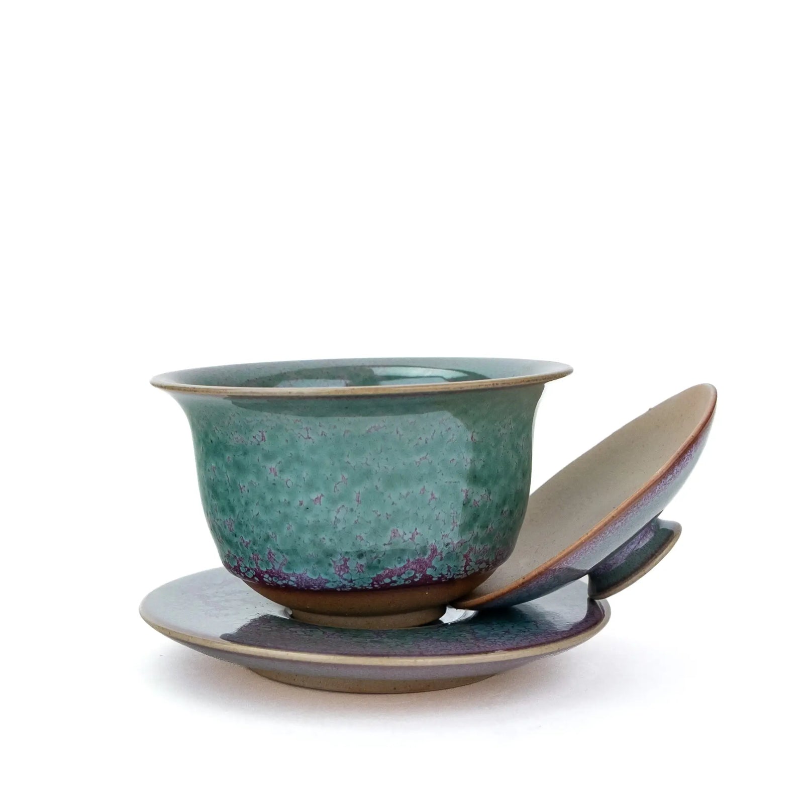 A handmade gaiwan with a matching saucer, featuring a speckled teal and purple glaze against a plain white background and the lid off