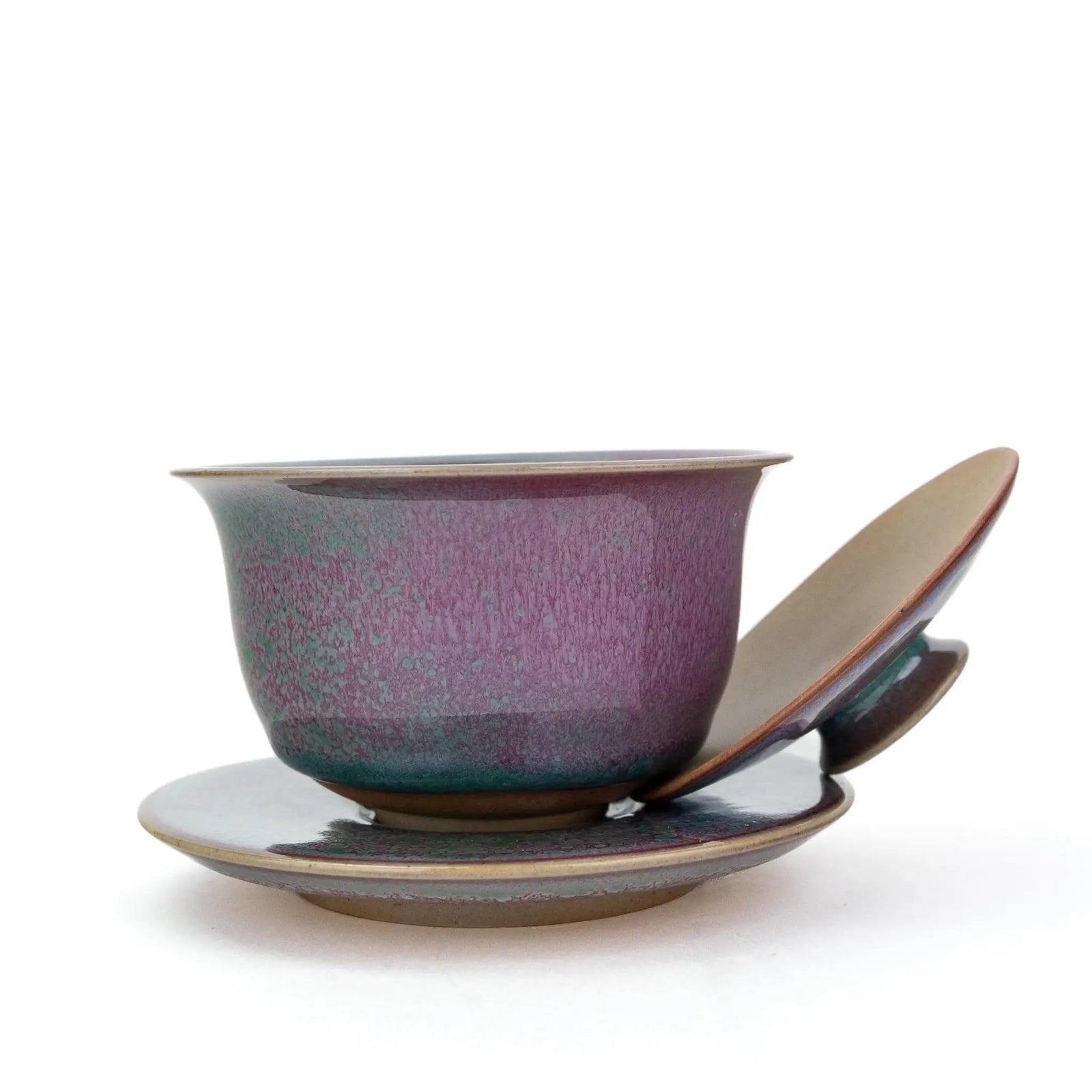 A ceramic handmade gaiwan with a matching saucer and lid, featuring a textured glaze that transitions from deep purple to teal green, with a warm brown rim, set against a plain white background.