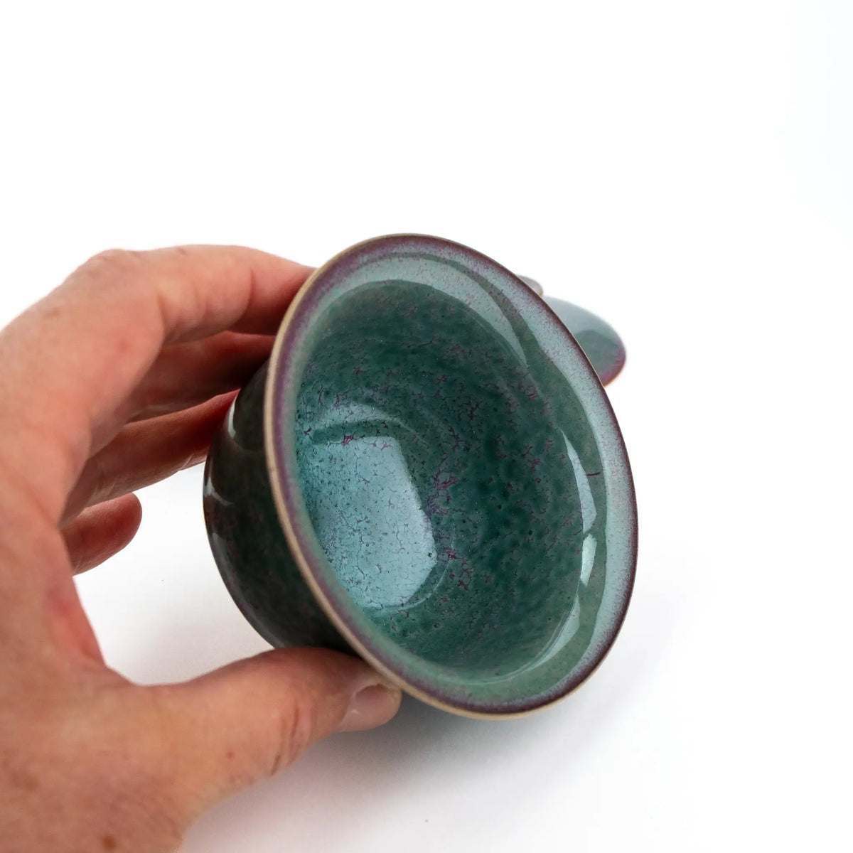 A hand holding a small ceramic cup with a glossy teal-green interior, shown against a plain white background.