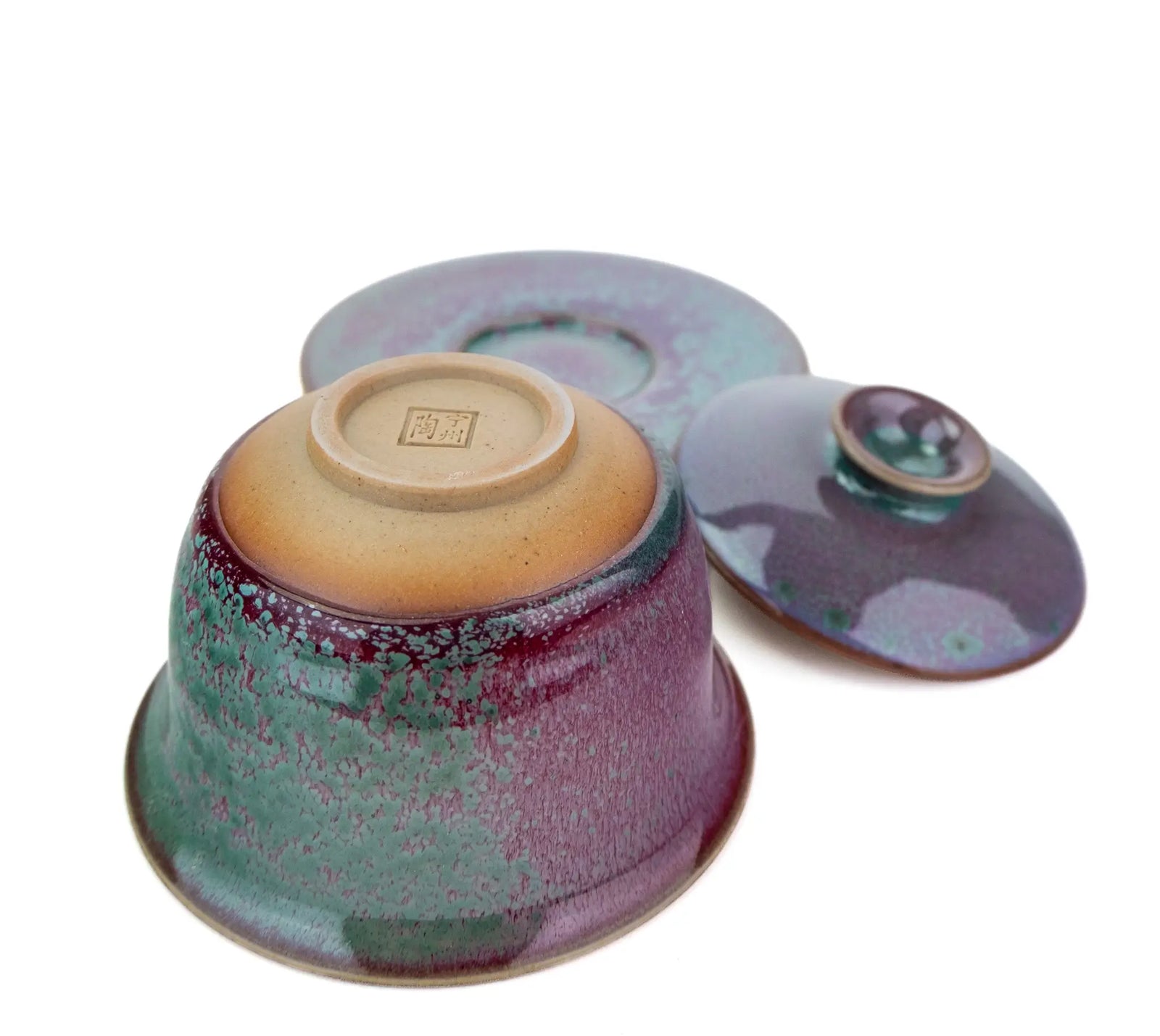 A ceramic handmade gaiwan shown upside down with its matching lid behind it, featuring a glossy glaze with a gradient of teal green, purple, and warm beige at the base.