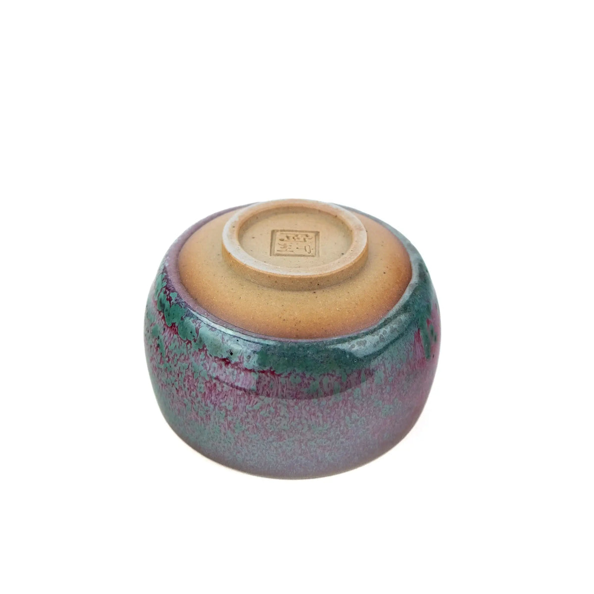 A handmade ceramic teacup with a glossy glaze featuring a gradient of purple, pink, and teal-green, with a natural brown clay base, set against a plain white background.  The view is of the underside. 