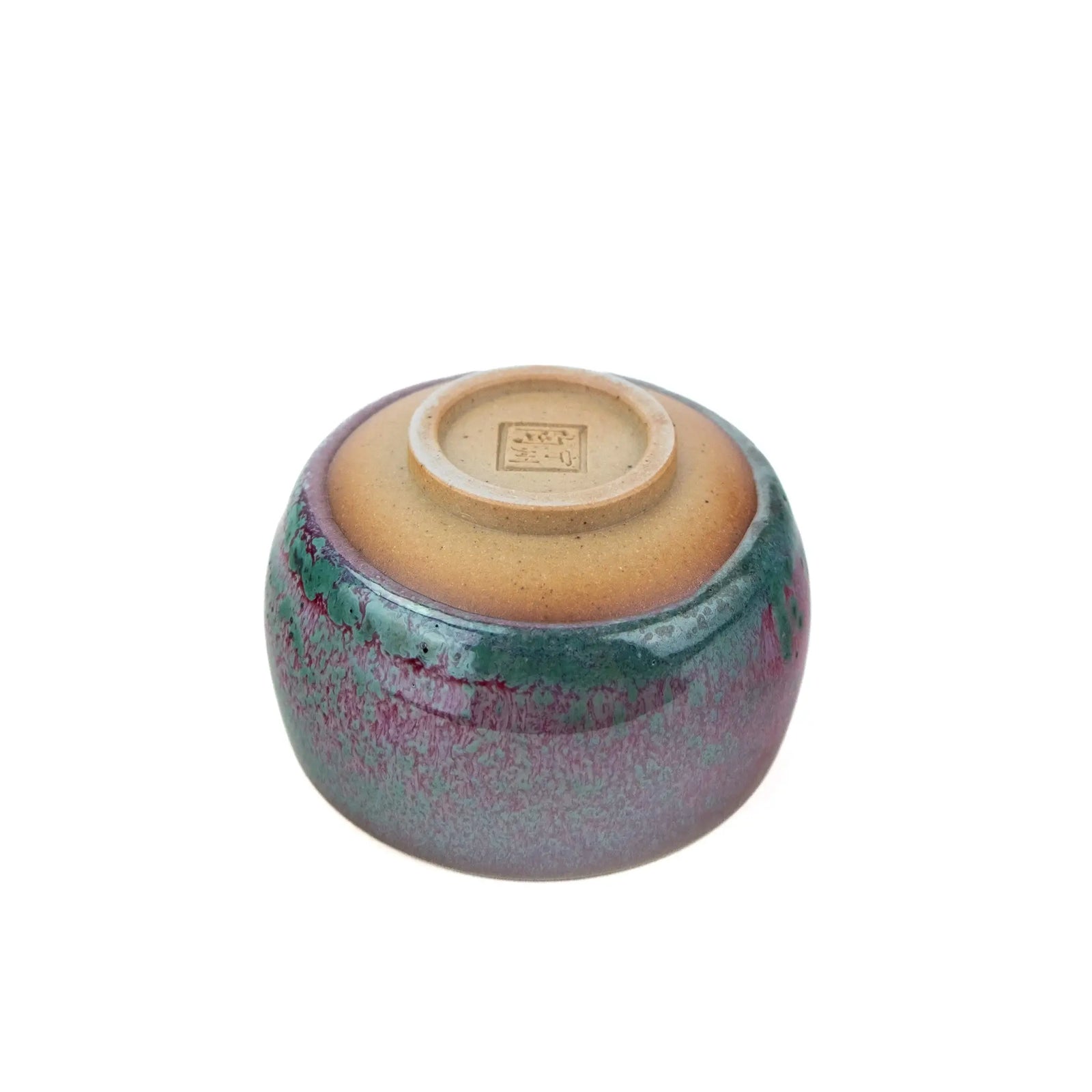 A handmade ceramic teacup with a glossy glaze featuring a gradient of purple, pink, and teal-green, with a natural brown clay base, set against a plain white background.  The view is of the underside. 